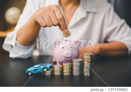 Hand putting coin into pink piggy bank beside toy car and stacked coins, showing car savings, down payment goals, auto loan budgeting and smart personal finance planning for vehicle purchase. Hand putting coin into pink piggy bank beside toy car and stacked coins, showing car savings, down payment goals, auto loan budgeting and smart personal finance planning for vehicle purchase. 135749062
