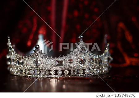 White crown with crystals, diamonds on dark red sparkle background. 135749276