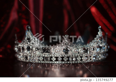 White crown with crystals, diamonds on dark red sparkle background. 135749277