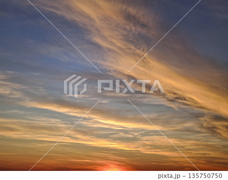The sun final rays cast a fiery red and orange glow at horizon. Wispy cirrus and altocumulus clouds stretch across a deep blue and gold sky, creating a painterly, textured effect of tranquil beauty 135750750
