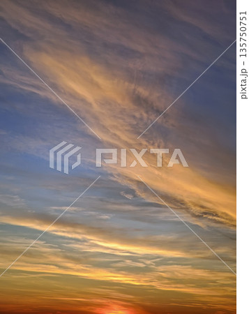 The sun final rays cast a fiery red and orange glow at horizon. Wispy cirrus and altocumulus clouds stretch across a deep blue and gold sky, creating a painterly, textured effect of tranquil beauty The sun final rays cast a fiery red and orange glow at horizon. Wispy cirrus and altocumulus clouds stretch across a deep blue and gold sky, creating a painterly, textured effect of tranquil beauty 135750751
