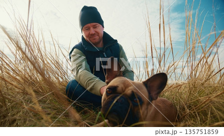A Person Enjoys a Relaxing Moment with a French Bulldog Amidst Tall Grass Under a Beautiful Sky, Emphasizing the Connection Between Humans and Their Pets in Nature's Embrace 135751859