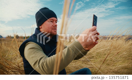 A Man Sitting in a Field Captures Nature's Beauty on His Smartphone, Embracing the Serenity of the Great Outdoors as He Enjoys a Peaceful Moment Surrounded by Tall Grass and a Clear Sky A Man Sitting in a Field Captures Nature's Beauty on His Smartphone, Embracing the Serenity of the Great Outdoors as He Enjoys a Peaceful Moment Surrounded by Tall Grass and a Clear Sky 135751871