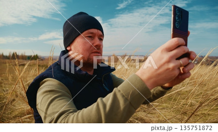 A Man in a Beanie Capturing a Photo with His Smartphone Amidst Tall Grass Under a Clear Blue Sky, Enjoying Nature and the Outdoors in a Peaceful Setting A Man in a Beanie Capturing a Photo with His Smartphone Amidst Tall Grass Under a Clear Blue Sky, Enjoying Nature and the Outdoors in a Peaceful Setting 135751872