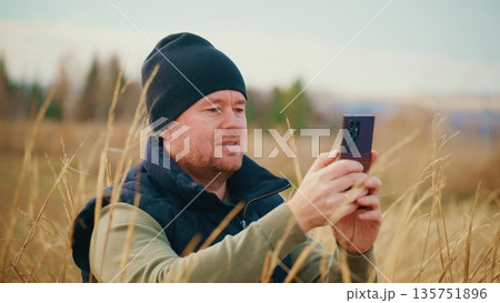 Man in Field Using Smartphone to Capture Nature's Beauty, Surrounded by Tall Grasses and Scenic Landscape Under Clear Sky, Enjoying Outdoor Photography Experience and Connection Man in Field Using Smartphone to Capture Nature's Beauty, Surrounded by Tall Grasses and Scenic Landscape Under Clear Sky, Enjoying Outdoor Photography Experience and Connection 135751896
