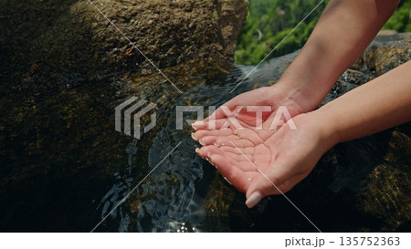 Human hands cupping clear spring water over natural stone basin in green forest setting creating peaceful outdoor wellness scene with gentle sunlight and fresh stream atmosphere 135752363