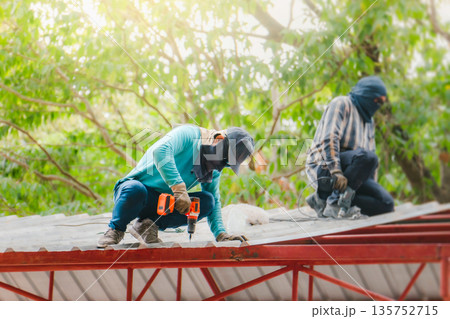 Roofer working in special protective work wear gloves, using air or pneumatic nail gun installing asphalt shingle on top of the new roof, Concept of residential building under construction. Roofer working in special protective work wear gloves, using air or pneumatic nail gun installing asphalt shingle on top of the new roof, Concept of residential building under construction. 135752715