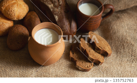A cozy food set with fresh milk in earthenware jug and mug, various types of artisan bread, including dark rye loaf and wheat rolls on a textured burlap fabric background. 135752763