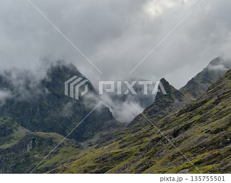 Majestic mountains rise through thick mist, their rugged peaks partially hidden by low clouds. Steep, green-tinted rocky slopes dominate the foreground on an overcast day. Majestic mountains rise through thick mist, their rugged peaks partially hidden by low clouds. Steep, green-tinted rocky slopes dominate the foreground on an overcast day. 135755501