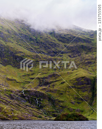 A majestic green mountain dominates the scene, partially covered by mist, with several narrow waterfalls flowing down its slopes into a calm lake. A small island sits in the water. 135755505