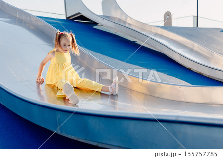 Little girl rides on playground slide, enjoying sunny summer day outdoors. 135757785