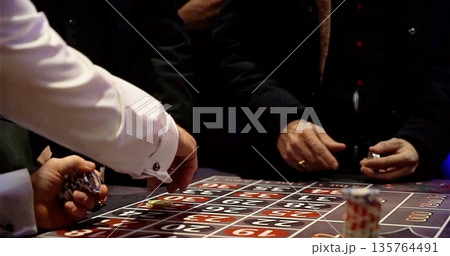 Gambling Game Scene, Individuals Strategically Placing Chips Around Roulette Table On Lowlit Gambling Floor 135764491