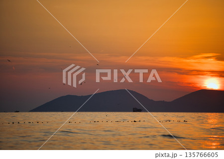 Vlora, Albania August , 2025 A serene sunset over the Adriatic Sea, with warm golden light reflecting across gentle waves and distant silhouettes of mountains. The dramatic clouds and soft evenin Vlora, Albania August , 2025 A serene sunset over the Adriatic Sea, with warm golden light reflecting across gentle waves and distant silhouettes of mountains. The dramatic clouds and soft evenin 135766605