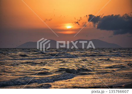 Vlora, Albania August , 2025 A serene sunset over the Adriatic Sea, with warm golden light reflecting across gentle waves and distant silhouettes of mountains. The dramatic clouds and soft evenin Vlora, Albania August , 2025 A serene sunset over the Adriatic Sea, with warm golden light reflecting across gentle waves and distant silhouettes of mountains. The dramatic clouds and soft evenin 135766607