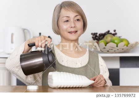 キッチンで湯たんぽにお湯を注ぐシニア女性 135769972
