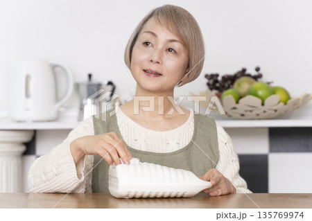 キッチンで湯たんぽにお湯を注ぐシニア女性 135769974