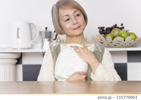 キッチンで湯たんぽにお湯を注ぐシニア女性 135770001