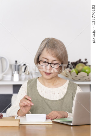 キッチンで家計簿をつけるシニア女性 キッチンで家計簿をつけるシニア女性 135770102
