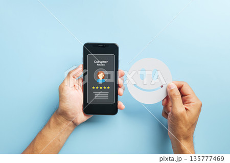 Review online customer feedback, hand holds smartphone displaying five star rating for positive service experience, another hand holds smiling face indicating high satisfaction Review online customer feedback, hand holds smartphone displaying five star rating for positive service experience, another hand holds smiling face indicating high satisfaction 135777469