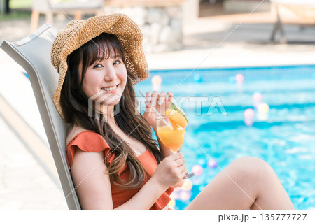 プールサイドのリクライニングチェアに座りトロピカルジュースを飲む水着姿の若いアジア人女性 プールサイドのリクライニングチェアに座りトロピカルジュースを飲む水着姿の若いアジア人女性 135777727