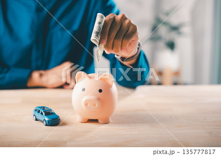 Man puts money in piggy bank to save on car fuel cost, gas price, insurance, auto finance, tax, saving funds for vehicle maintenance, travel, and road trip transportation expenses Man puts money in piggy bank to save on car fuel cost, gas price, insurance, auto finance, tax, saving funds for vehicle maintenance, travel, and road trip transportation expenses 135777817