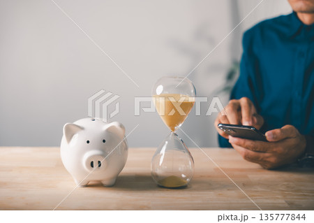 Hourglass and piggy bank help company save money, cost saving business budget and account time while man uses smartphone for online investment financial productivity on wooden table Hourglass and piggy bank help company save money, cost saving business budget and account time while man uses smartphone for online investment financial productivity on wooden table 135777844