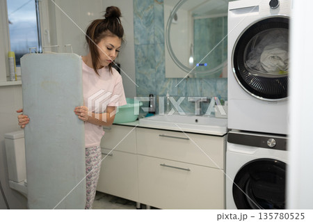Woman Holding Ironing Board in Bathroom Next to Stacked Washer and Dryer Woman Holding Ironing Board in Bathroom Next to Stacked Washer and Dryer 135780525