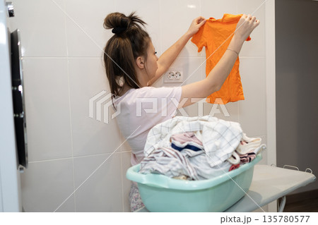 Woman Sorts Laundry Near Washing Machine in Bright Tiled Bathroom 135780577