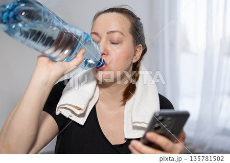 Woman Drinks Water While Using Phone in Indoor Space After Workout Session Woman Drinks Water While Using Phone in Indoor Space After Workout Session 135781502