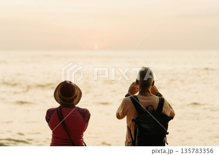 An adult couple of tourists, husband and wife, meet the sunset at sea. They look at the setting sun and take pictures on their smartphone, rear view. High quality photo An adult couple of tourists, husband and wife, meet the sunset at sea. They look at the setting sun and take pictures on their smartphone, rear view. High quality photo 135783356