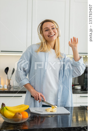 Young woman slicing banana for smoothie blender while dancing with headphones for healthy fit diet Young woman slicing banana for smoothie blender while dancing with headphones for healthy fit diet 135783407