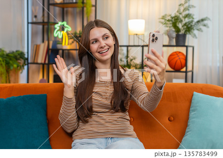 Young woman at home starts video call smiles looks at smartphone camera makes greetings hello sign Young woman at home starts video call smiles looks at smartphone camera makes greetings hello sign 135783499