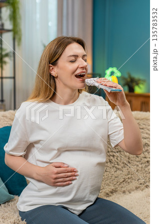 Pregnant woman eats dark chocolate rubs belly sweet happy relax moment cocoa treat on sofa at home 135783532