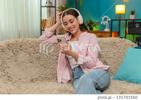 Young woman listening to podcast on sofa wearing headphones nodding while writing insightful notes Young woman listening to podcast on sofa wearing headphones nodding while writing insightful notes 135783560