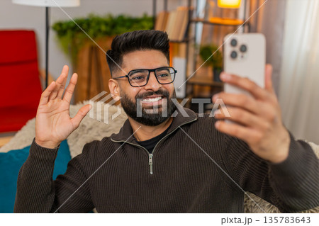 Indian man filming selfie video on smartphone at home, smiling and waving to followers cheerfully Indian man filming selfie video on smartphone at home, smiling and waving to followers cheerfully 135783643