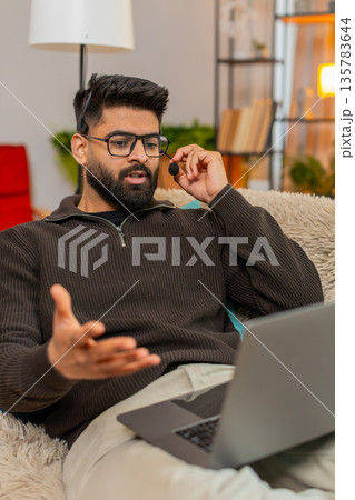 Indian man using wireless headset speaks on laptop video call with client while working at home Indian man using wireless headset speaks on laptop video call with client while working at home 135783644