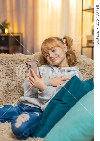Young happy teenager girl looking at family photos on smartphone at home sofa laugh, finger pointing 135783798