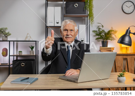 Happy Caucasian elderly businessman using laptop and showing thumbs up at camera in home office 135783854