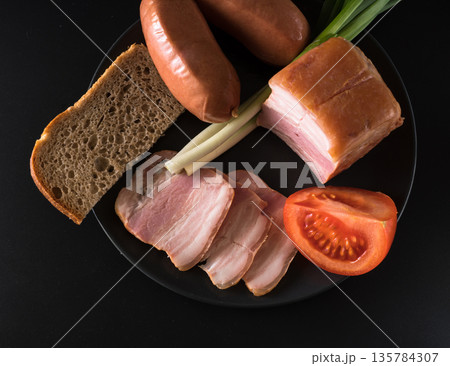 Food, lard, green onions, tomato, black bread, sausages, on a black plate on a black background Food, lard, green onions, tomato, black bread, sausages, on a black plate on a black background 135784307