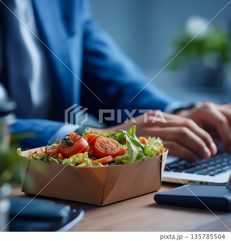 Employee enjoying healthy office lunch from takeaway box. Businessman working at desk with fresh salad meal in sustainable paper lunchbox Employee enjoying healthy office lunch from takeaway box. Businessman working at desk with fresh salad meal in sustainable paper lunchbox 135785504