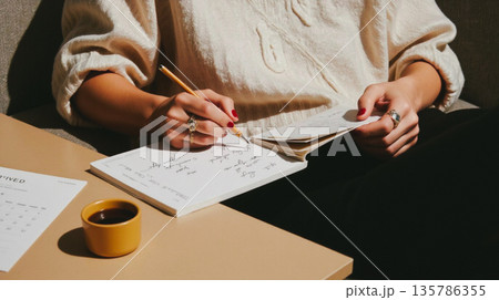 A cozy, authentic lifestyle shot of a woman in a fluffy cream sweater writing in a blank open notebook or planner, diary with a pen. Perfect for self-care, wellness, and creative planning concepts.  135786355