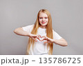 Happy young girl model with long blond hair making heart gesture with hands, smiling in studio, isolated on grey wall background in bright positive portrait 135786725