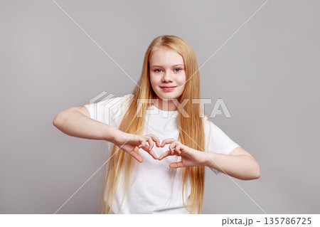 Happy young girl model with long blond hair making heart gesture with hands, smiling in studio, isolated on grey wall background in bright positive portrait 135786725