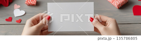 Woman's hand holding blank vertical card for Valentine's Day. Woman's hand holding white blank greeting card mockup with scattered red hearts 135786805