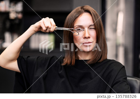 Beautiful female hairstylist looking through hairdressing scissors while sitting in modern hair salon. Professional woman in dark clothes, indoor beauty studio, creative portrait, fashion and style Beautiful female hairstylist looking through hairdressing scissors while sitting in modern hair salon. Professional woman in dark clothes, indoor beauty studio, creative portrait, fashion and style 135788326