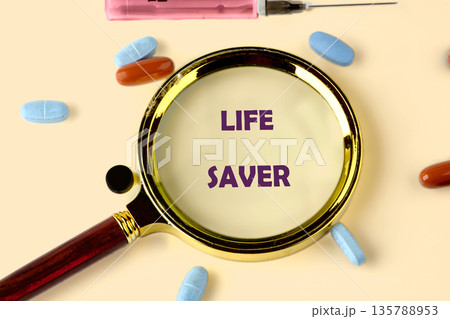 A magnifying glass focuses on the words LIFE SAVER surrounded by various pills and a syringe, emphasizing the importance of medicine and healthcare 135788953