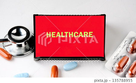 A stethoscope, pills and a sign with the text HEALTHCARE are arranged on a white surface. This evokes the theme of medical care and the provision of services A stethoscope, pills and a sign with the text HEALTHCARE are arranged on a white surface. This evokes the theme of medical care and the provision of services 135788955