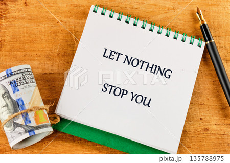 on a wooden surface, a notebook with the words LET NOTHING STOP YOU sits beside a stack of United States currency and an elegant fountain pen, inspiring ambition 135788975