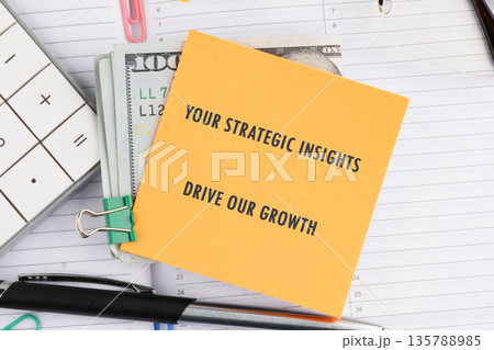 An orange sticky note proclaims YOUR STRATEGIC INSIGHTS DRIVE OUR GROWTH, resting atop a stack of United States dollar bills 135788985