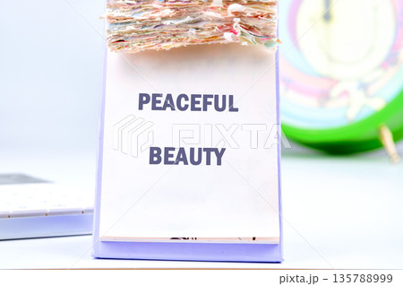 A crisp white page on a desktop calendar powerfully displays the uplifting words Peaceful Beauty. Many colorful, worn pages sit above, marking time 135788999